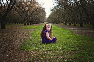 Young girl in a purple dress and floral headband sits on green grass between rows of bare trees in an orchard.
