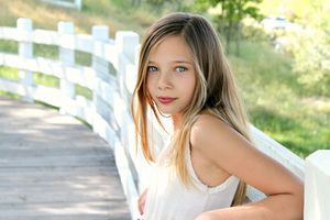 Young girl with long blonde hair and blue eyes leaning on a white wooden fence on a sunny wooden bridge.