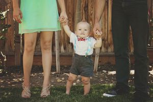 Toddler in a red bow tie and shorts taking steps while holding the hands of two parents standing on grass.