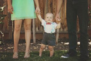 Baby in a bow tie and shorts takes standing steps while holding the hands of two parents on a grassy lawn.
