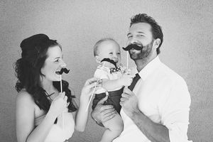 Black and white family portrait of a mother, father and baby holding playful paper mustaches on sticks.