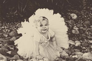 Smiling baby girl in a white tutu and flower headband sitting on rocks in a sepia toned portrait.