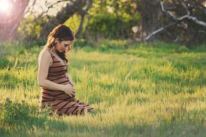Pregnant woman in a striped dress kneels in a golden sunlit grassy field and cradles her baby bump.