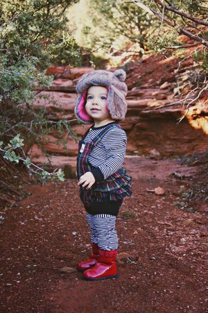 Toddler in a furry trapper hat, striped shirt, plaid overalls and red boots stands on a red dirt desert trail.