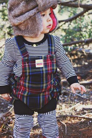 Close-up of a toddler in a fur trapper hat, striped top and plaid overalls standing outdoors among dry brush.