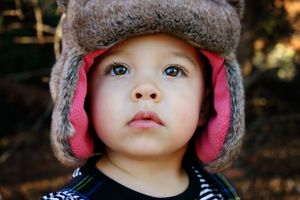 Close-up portrait of a wide-eyed toddler wearing a gray fur trapper hat with pink lining against a blurred woodland background.