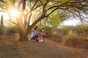 Mother and young child sitting together beneath a large desert tree in warm, glowing sunset light surrounded by tall grass.