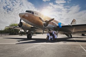 Group of people posing beneath the nose of a vintage propeller airplane on an airfield under a bright sunburst sky.