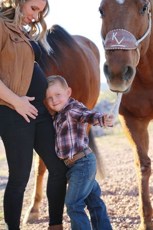 A smiling boy in a plaid shirt and jeans leans against his pregnant mother while holding the rope of a brown horse.
