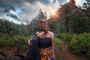 Two women in colorful African print outfits embrace in a green forest with red rock formations and a glowing sunset behind them.
