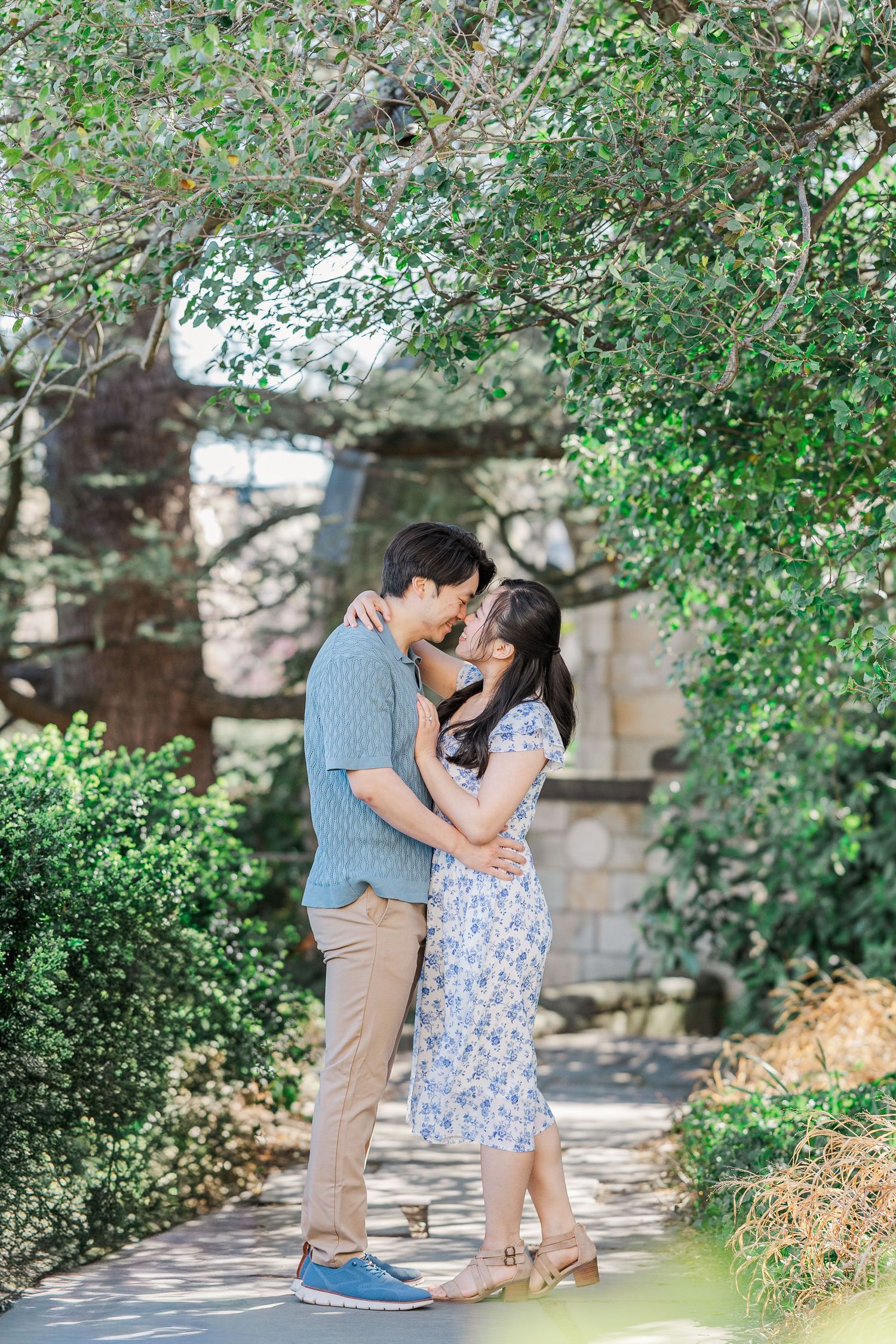 A couple shares a romantic kiss surrounded by lush greenery in a sunlit garden pathway.