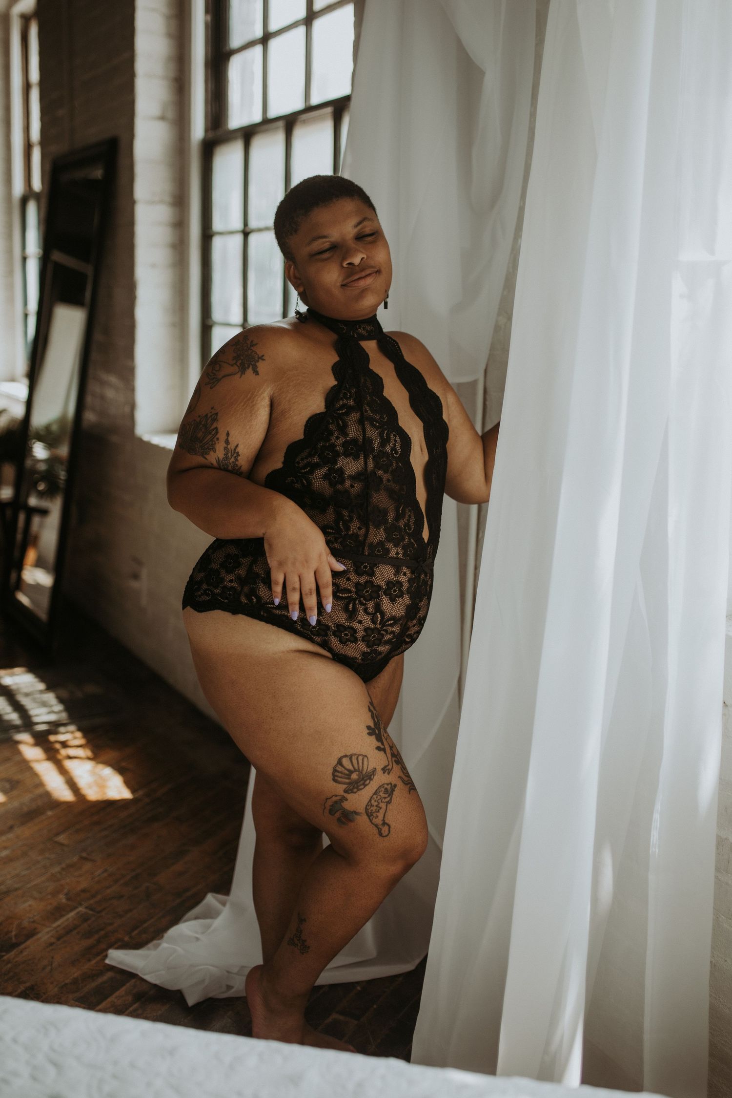 Plus-size model in black lace bodysuit with tattoos posing by sheer white curtains in natural light.