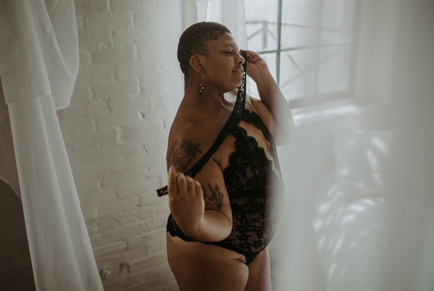 A plus-size woman in black lace lingerie poses gracefully near sheer curtains in a softly lit boudoir setting.