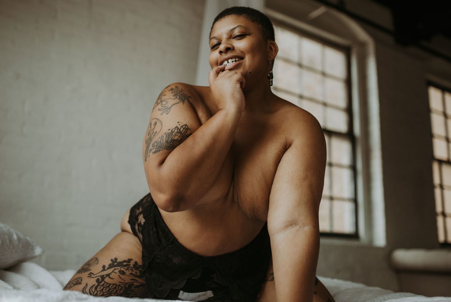 Confident plus-size woman with tattoos posing in black lace lingerie on a bed near a window.