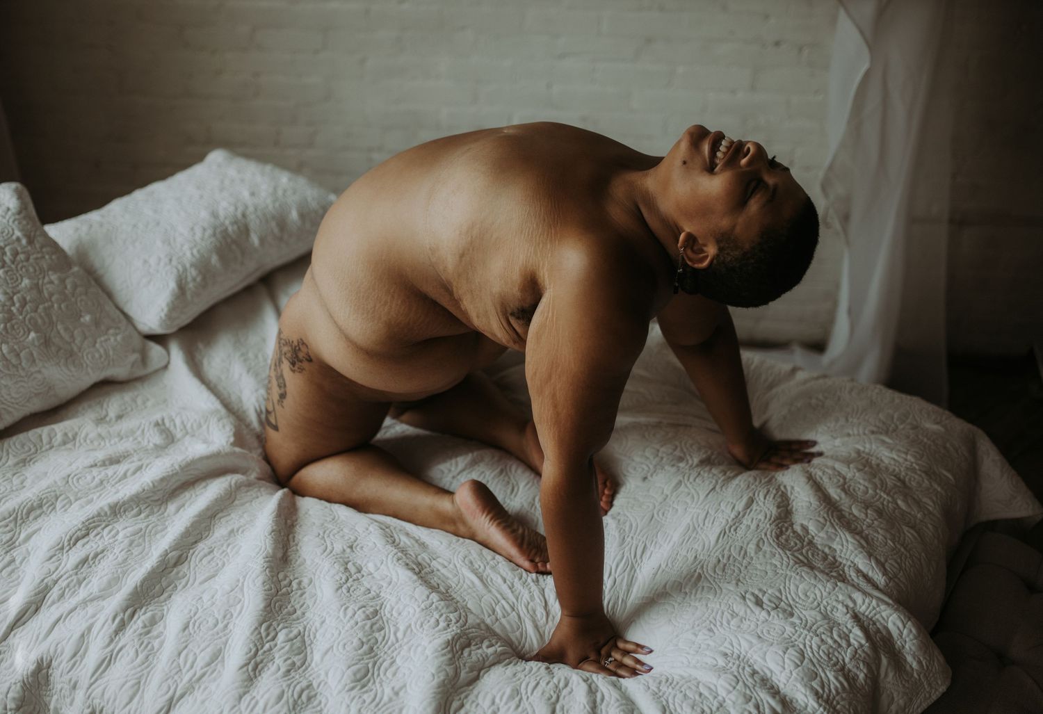A shirtless person kneeling on a white bed, leaning forward with head tilted back in a serene pose.