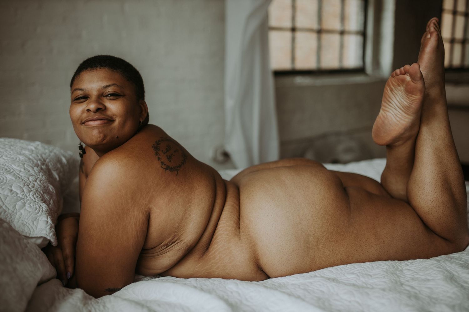 Plus-size person posing nude on a white bed, smiling confidently in a boudoir photography setting.