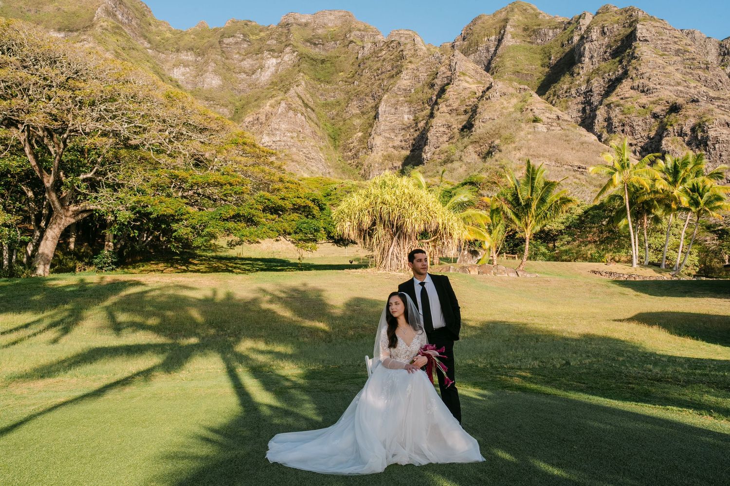Bride and groom pose on lush green lawn with dramatic Ko'olau mountain backdrop in Hawaii.