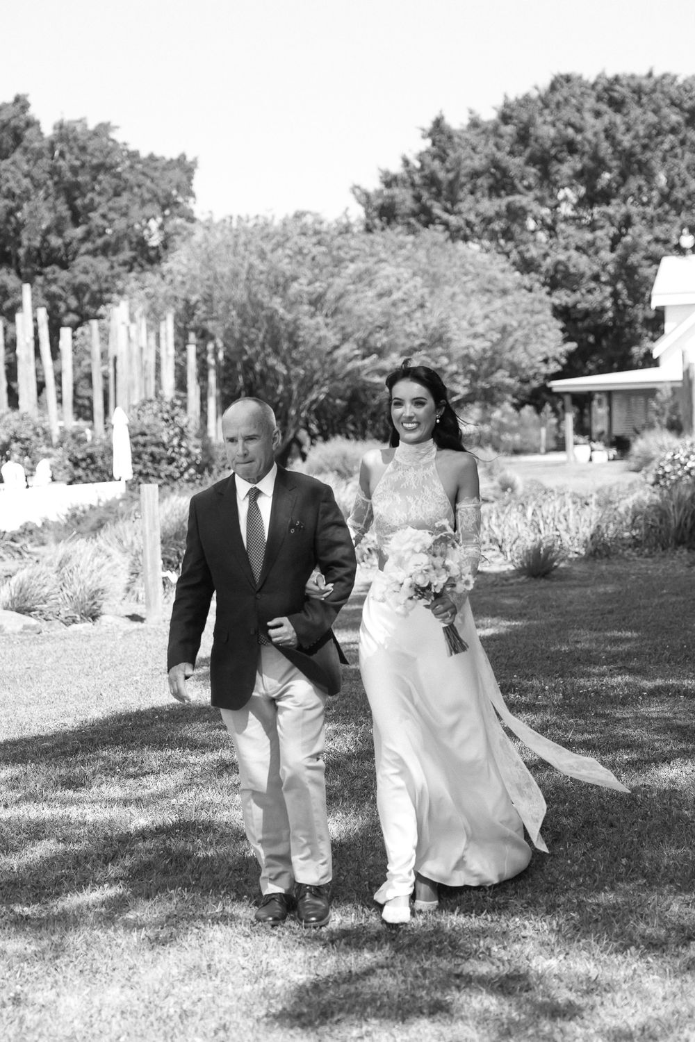 Black and white photo of a bride in a flowing gown walking with her father through a garden on her wedding day.