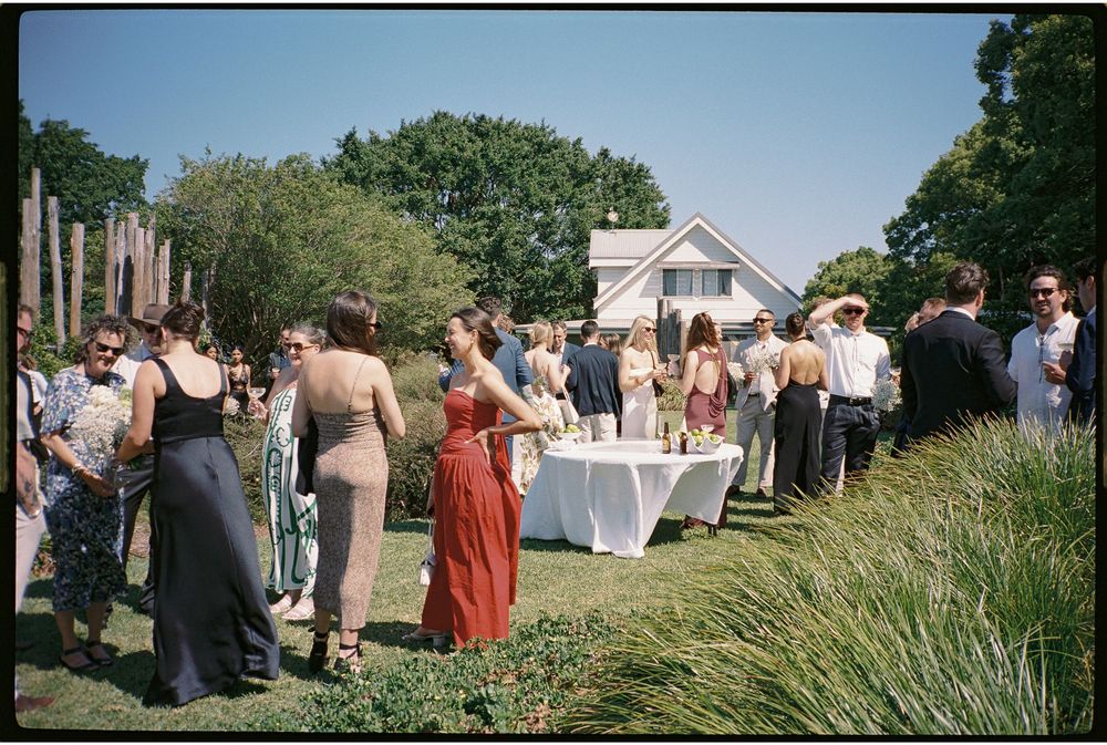 Guests mingling outdoors at a summer garden party near a white house, dressed in elegant attire on a sunny day.