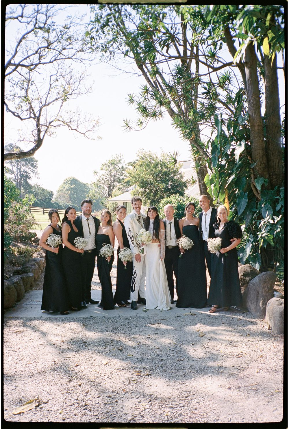 Wedding party in black attire with bride and groom posing outdoors on a gravel path surrounded by lush trees.