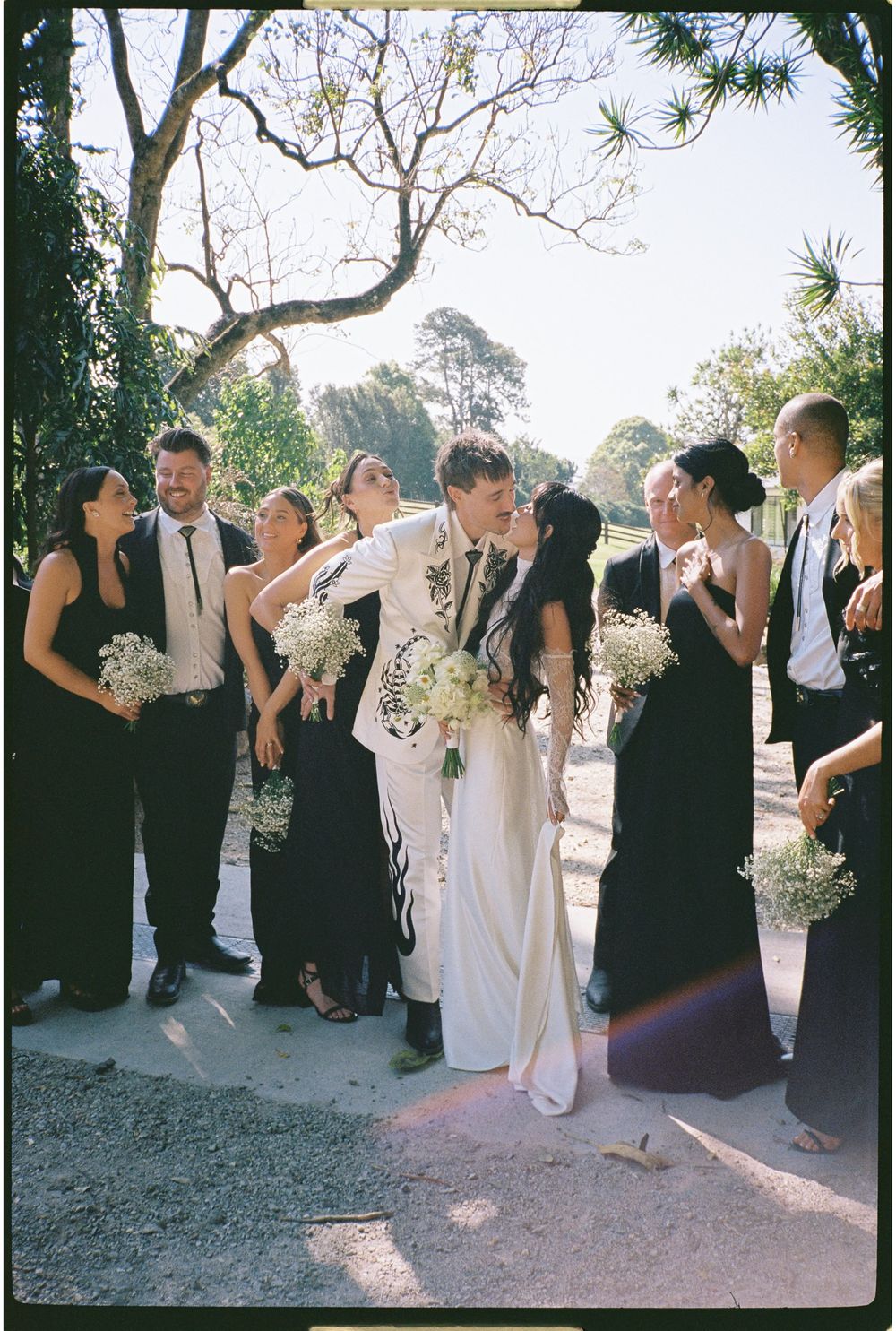 Bride and groom share a kiss surrounded by wedding party in black attire, holding white floral bouquets outdoors.