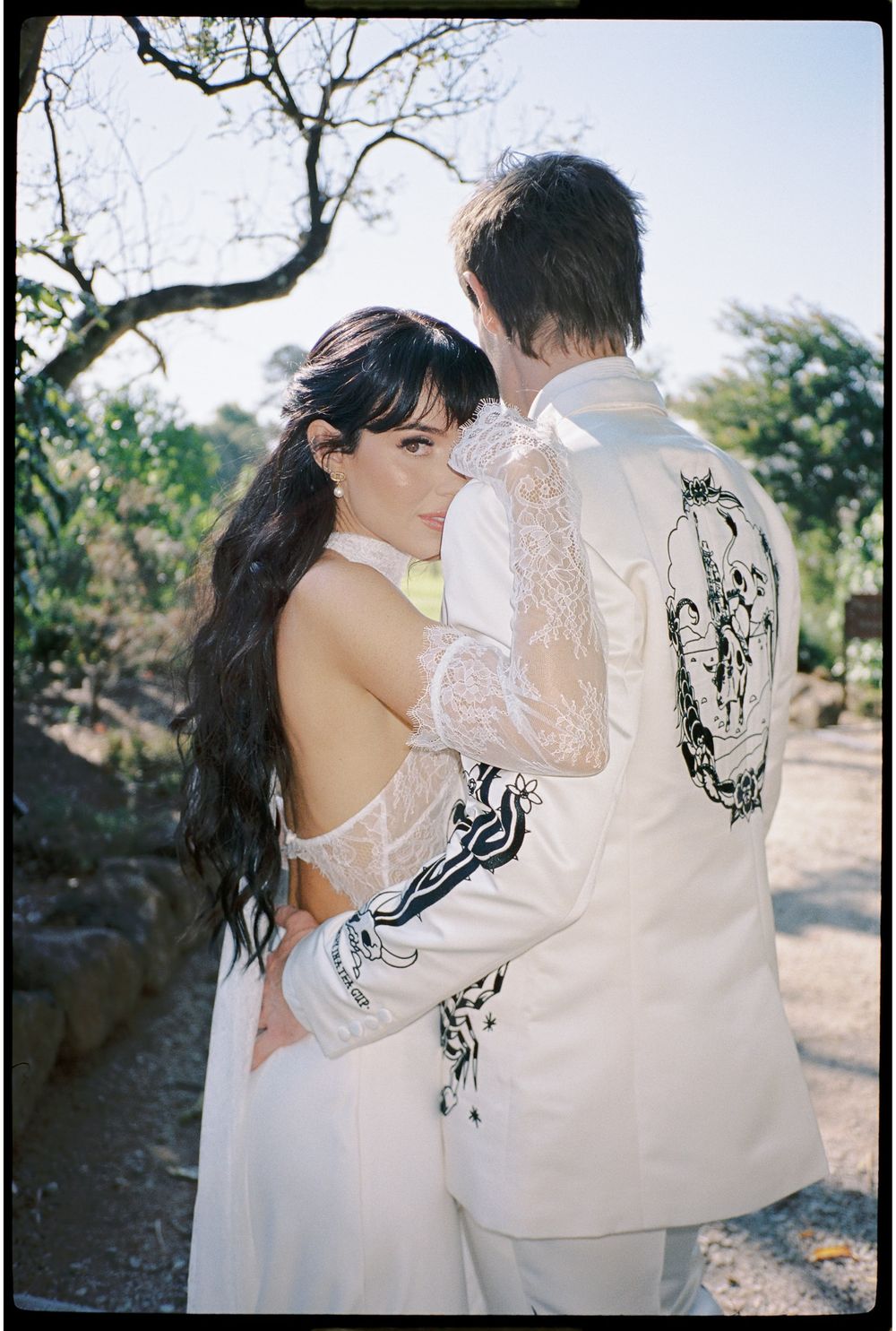 Couple embracing outdoors in wedding attire, woman in lace backless gown, man in embroidered white suit jacket.