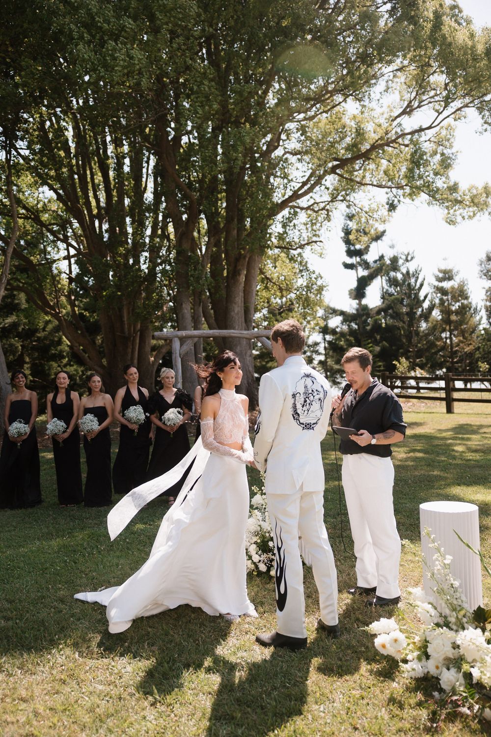 Outdoor wedding ceremony on green lawn with bride in white gown, groom, officiant, and bridesmaids in black dresses.