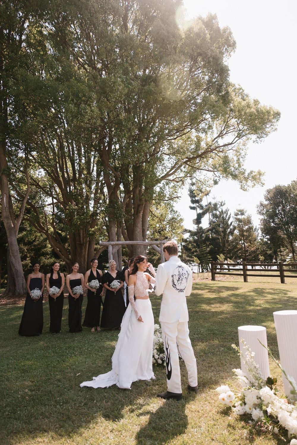 Outdoor wedding ceremony on green lawn with bride in white gown, groom, and bridesmaids in black dresses under tall trees.