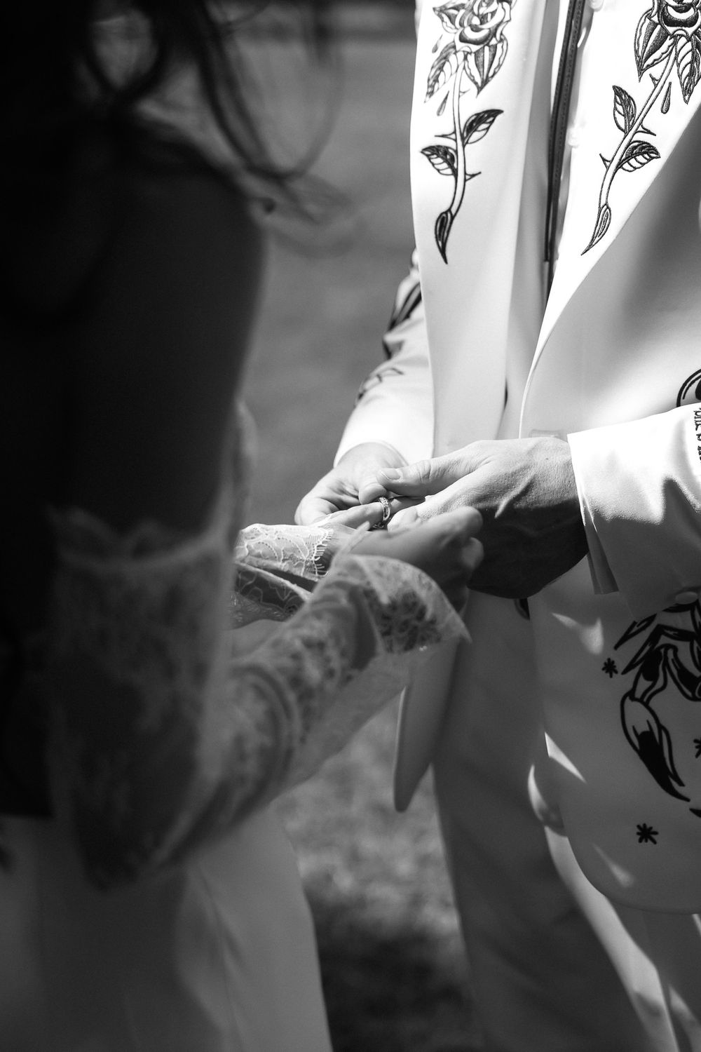 Black and white photo of a groom placing a wedding ring on the bride's tattooed hand during a ceremony.