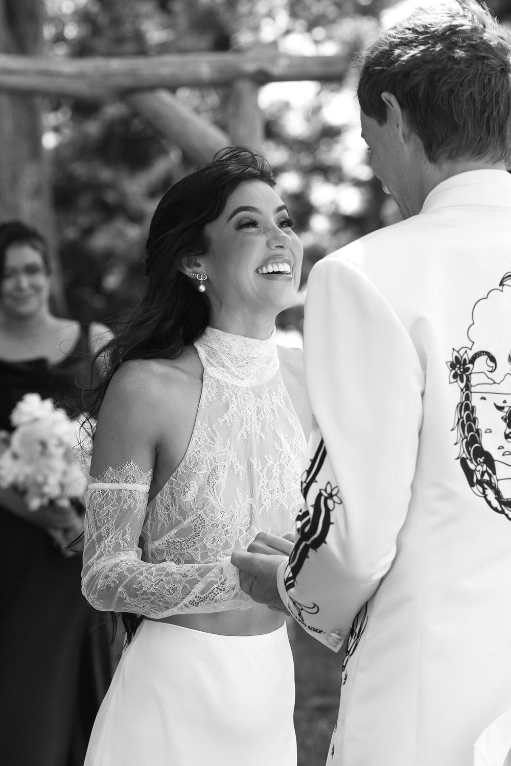 Black and white photo of a smiling bride in lace halter gown laughing with groom in embroidered white jacket at outdoor wedding.