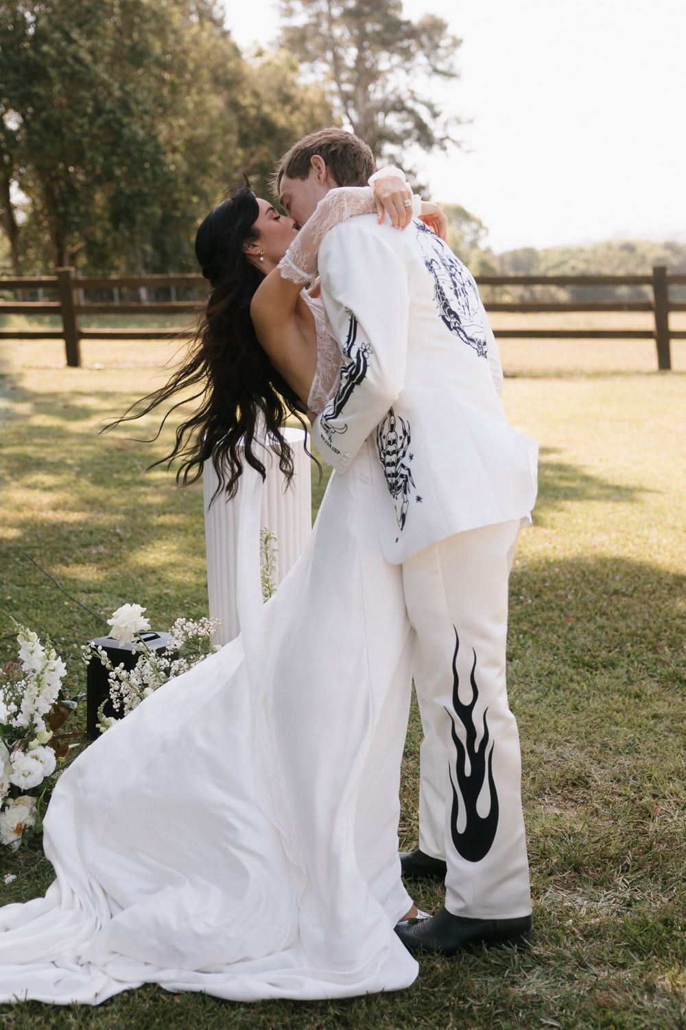 A bride and groom share a kiss outdoors, both wearing matching white outfits with black flame and tattoo designs at their wedding.
