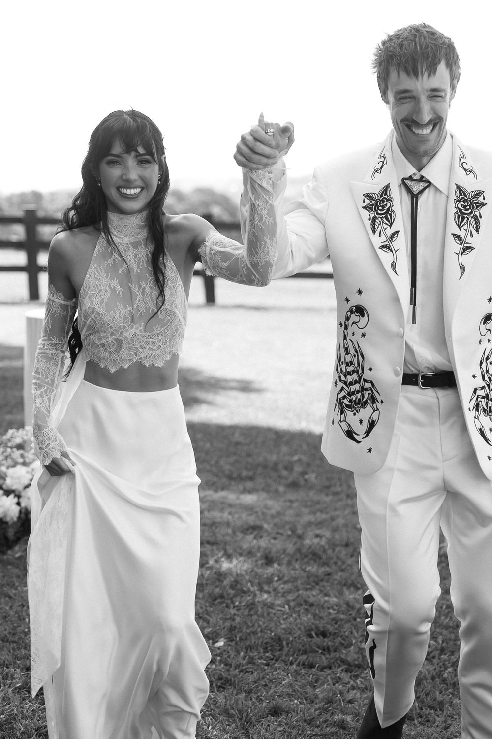 Black and white photo of smiling couple in wedding attire, groom in embroidered white suit, bride in lace crop top and skirt.