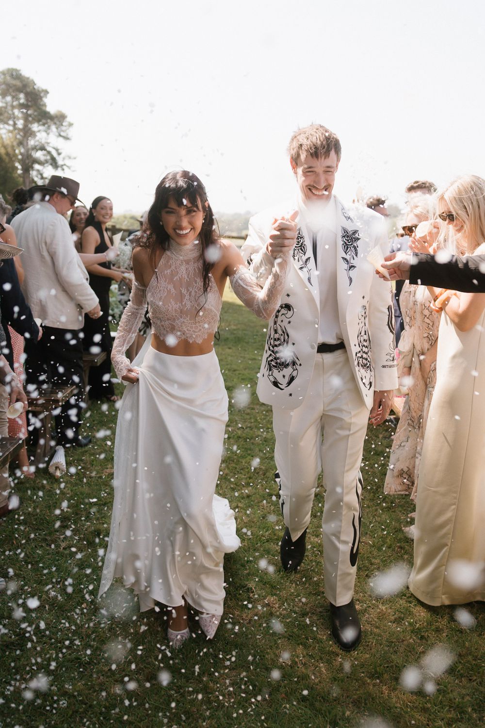 A joyful couple walks through a confetti shower at their outdoor wedding, bride in white crop top and groom in decorated suit.