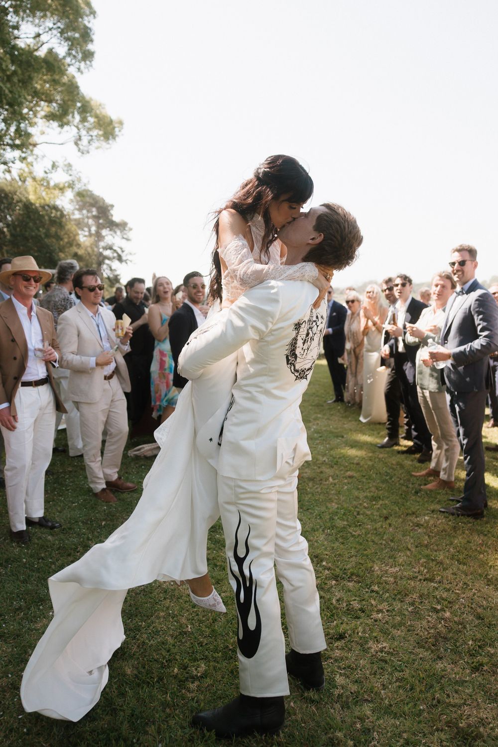 Groom in white flame suit lifts bride for a kiss outdoors, surrounded by wedding guests on a sunny lawn.