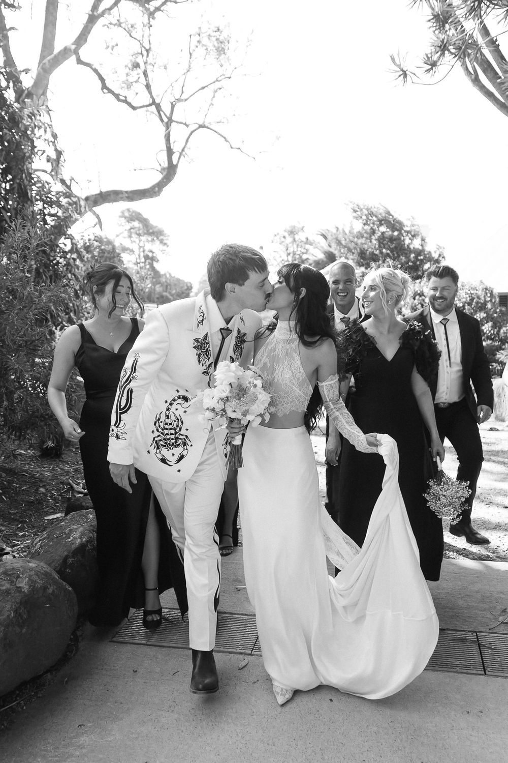 Black and white photo of a couple kissing on their wedding day, surrounded by bridesmaids and groomsmen walking outdoors.