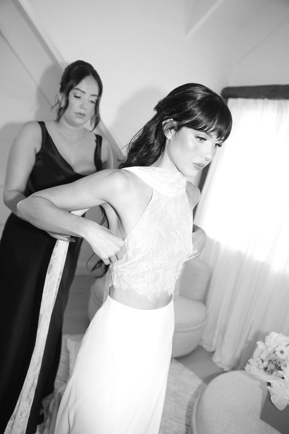 Black and white photo of a bride in a white two-piece gown being helped by a bridesmaid in a black dress.