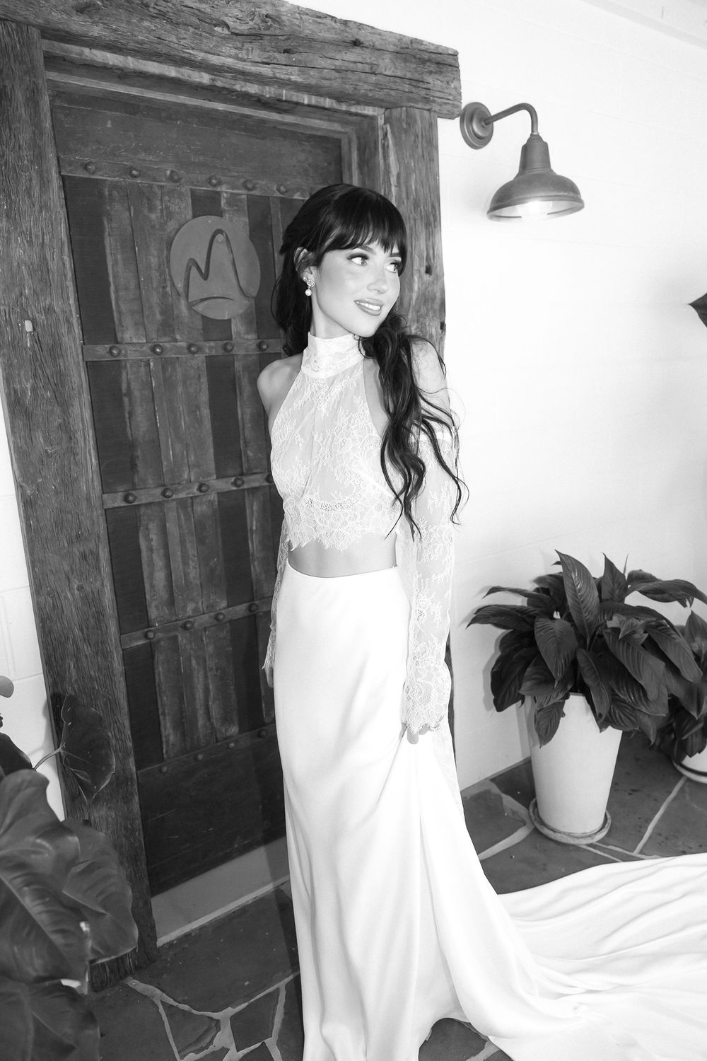 Black and white photo of a smiling woman in a white lace crop top and flowing skirt posing near a wooden door.