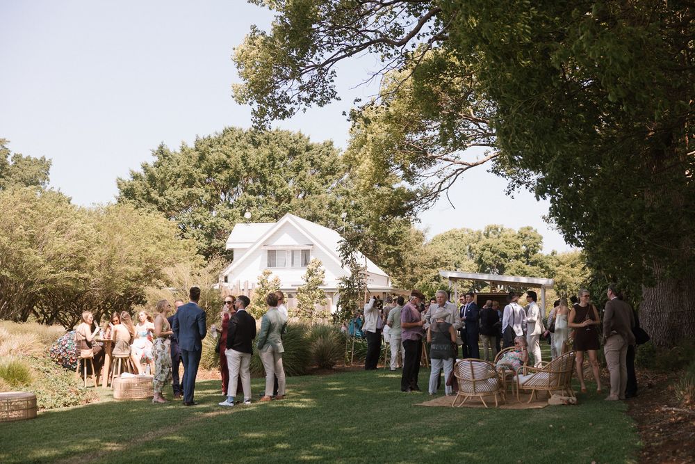 Guests mingle outdoors on a green lawn surrounded by trees near a white house during a garden party event.