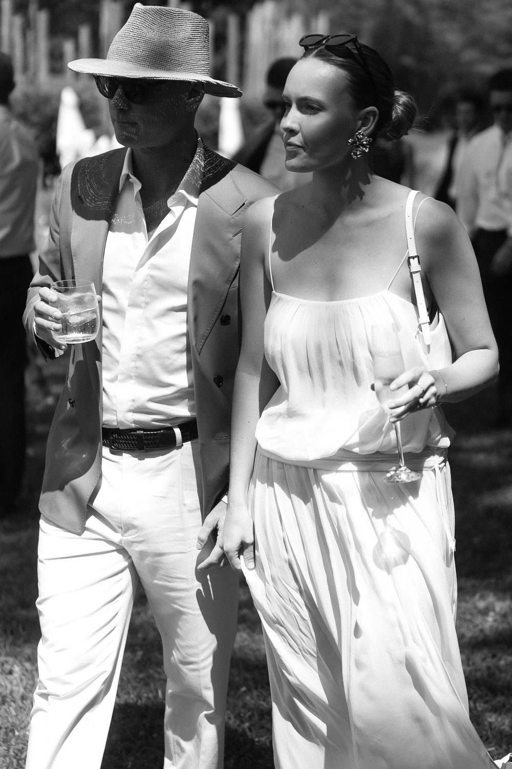 Elegant couple in white attire holding wine glasses at an outdoor summer event, black and white photography.