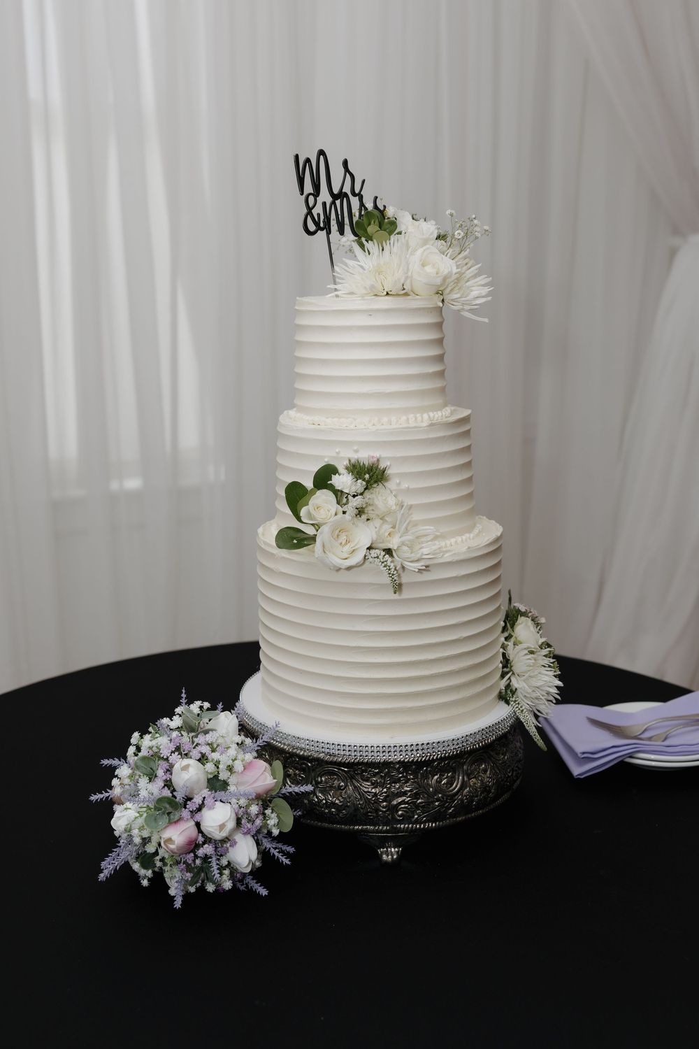 Elegant four-tier white wedding cake with ribbed frosting, white floral accents, and Mr. & Mrs. topper on silver stand.