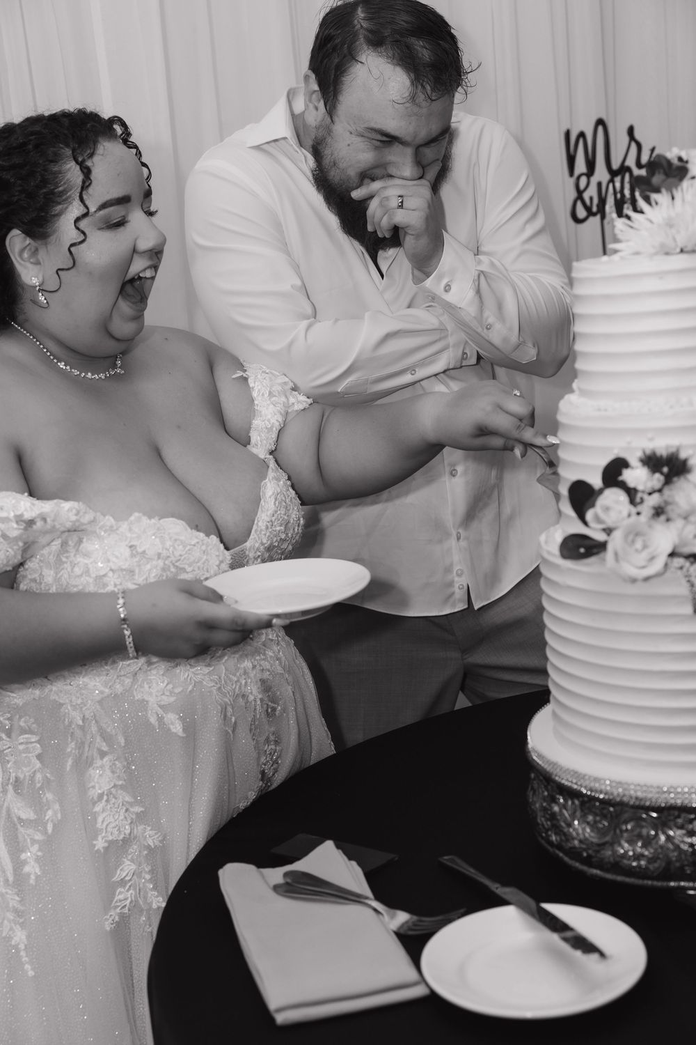 Black and white photo of a bride and groom laughing while cutting their wedding cake at their reception.