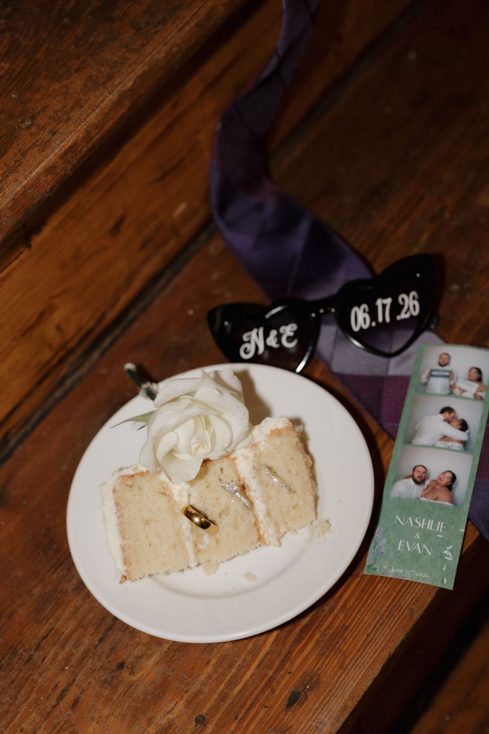 Wedding cake slice on white plate with gold rings, heart sunglasses, photo strip, and purple ribbon on wood steps.