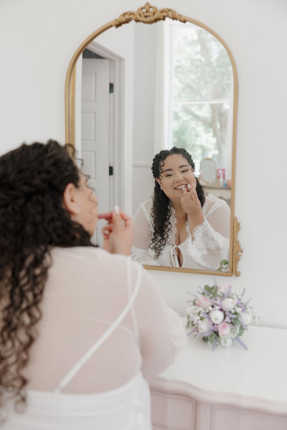 Bride applying lipstick while smiling at her reflection in an ornate gold mirror, with a floral bouquet nearby.