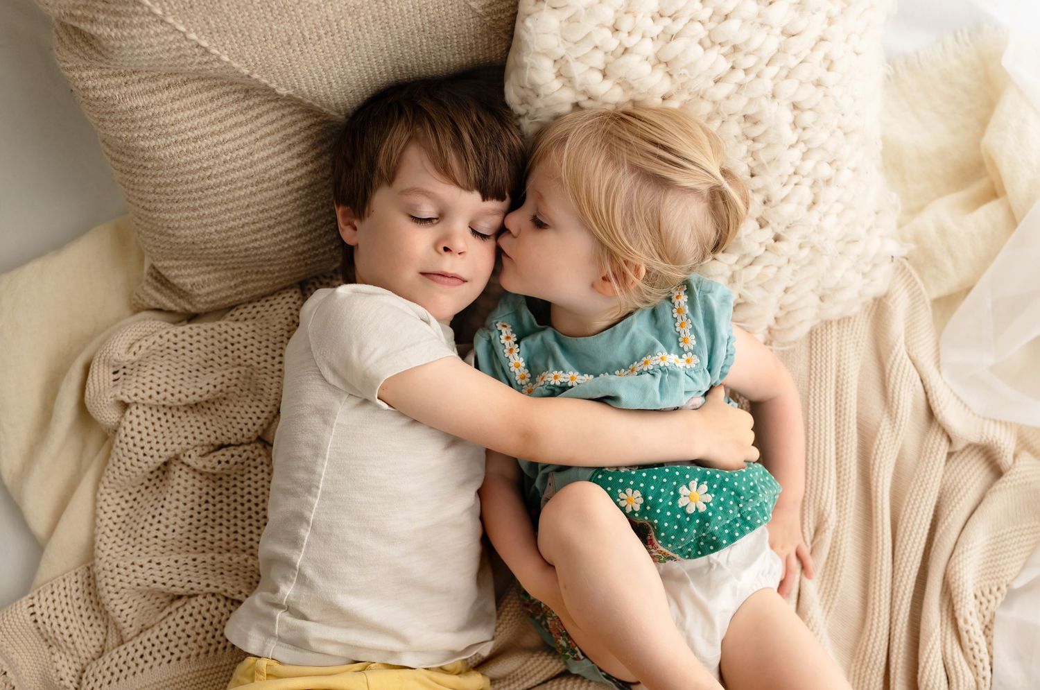 Blond toddler girl in blue kisses her brother's cheek as they lie down on soft, neutral sheets, by Charlottesville family photographer.