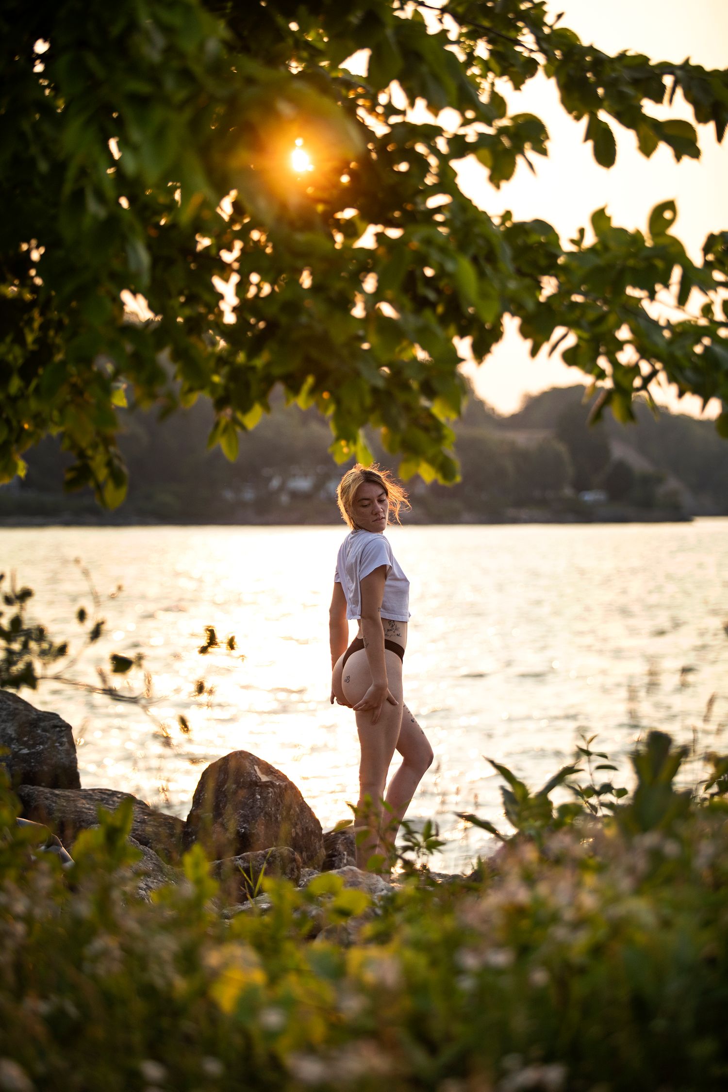 Sunset nature photo of someone standing on rocks by a lake with tree branches framing the scene.