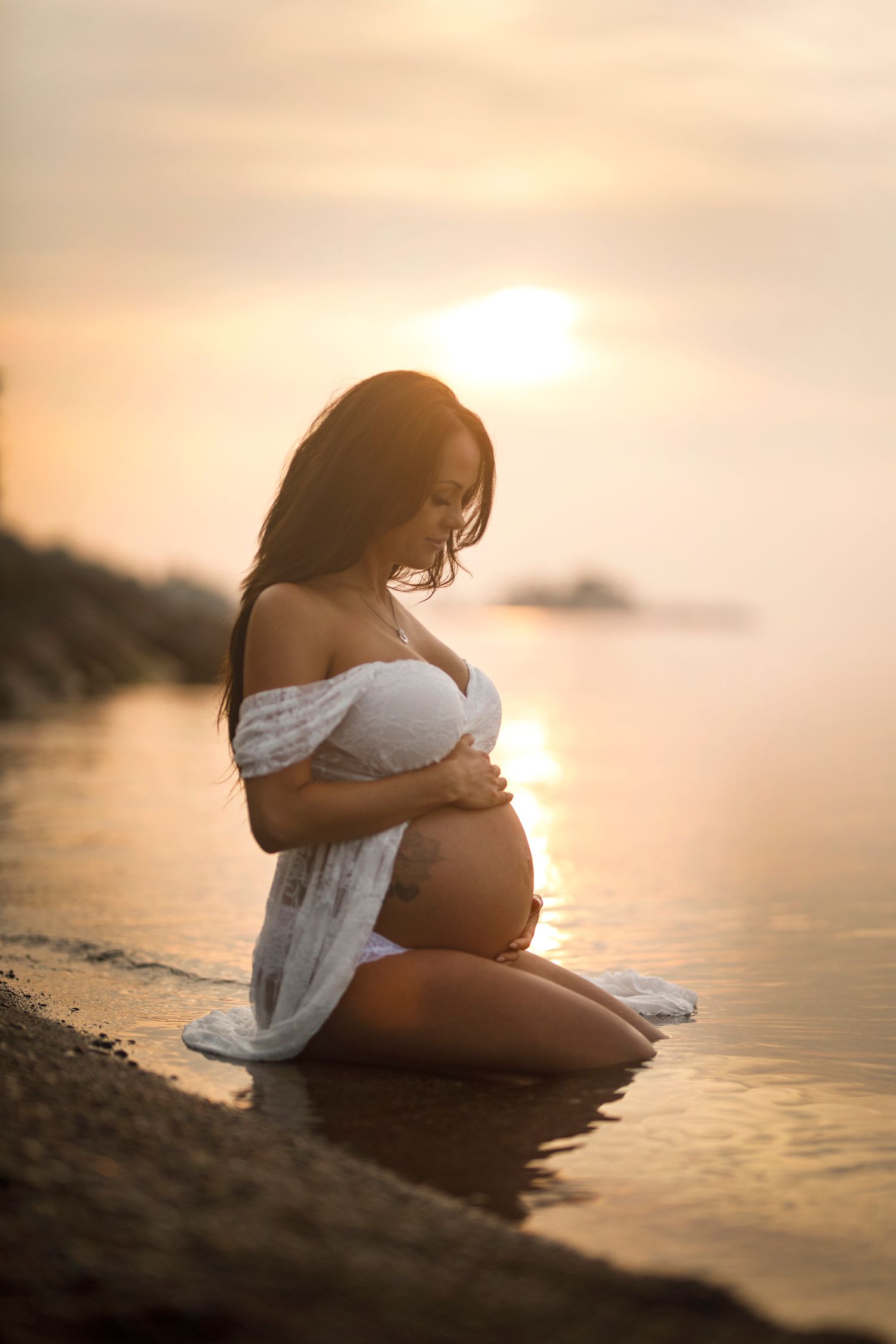 Pregnant woman in white bikini kneeling on beach at golden sunset, cradling her baby bump near calm ocean waters.