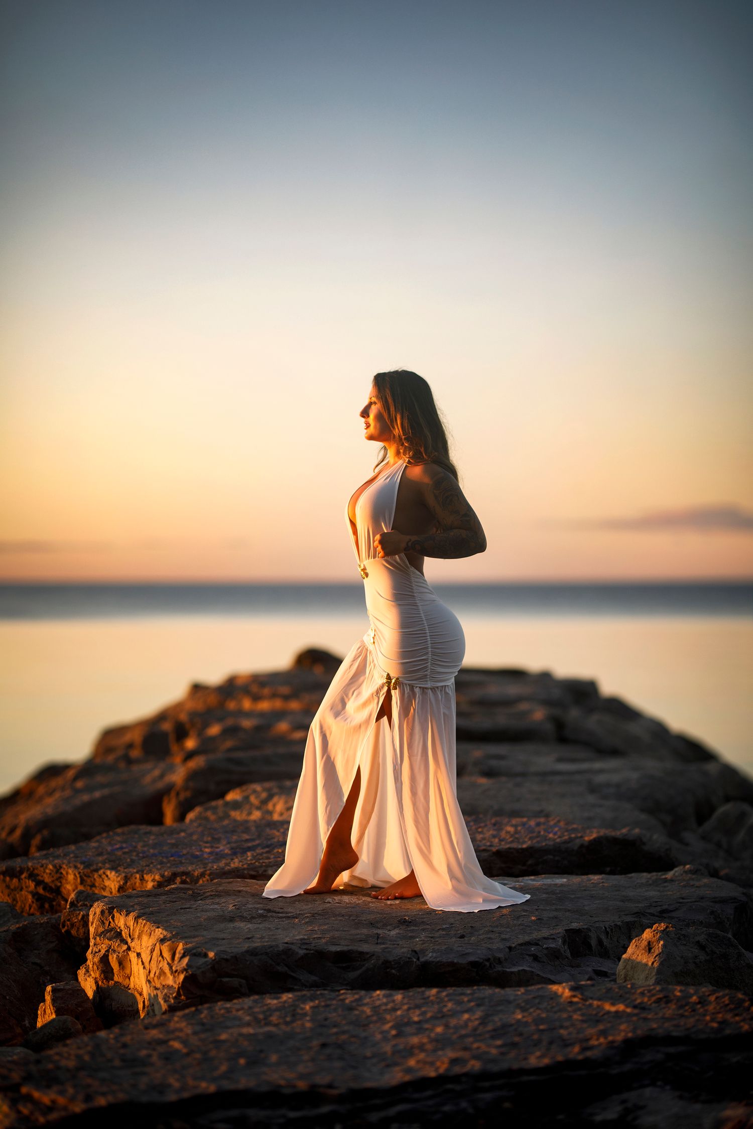 Woman in white dress standing on rocky pier at sunset with calm ocean and golden sky backdrop.