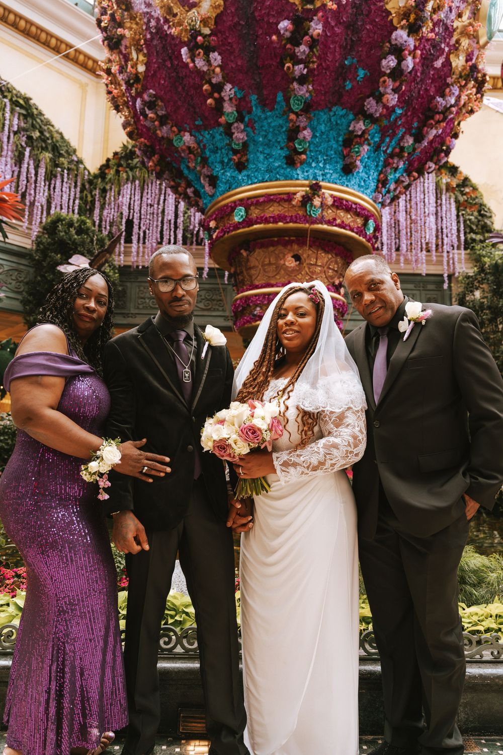 A bride in white lace gown holds bouquet posing with three guests near colorful floral fountain display.
