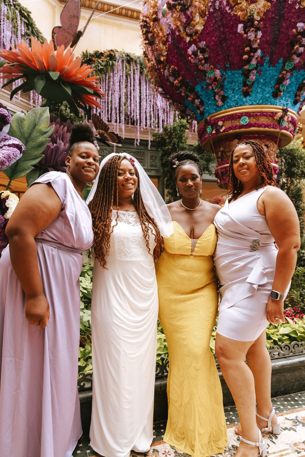 Bride in white gown and veil poses with three bridesmaids in colorful dresses amid floral decor.