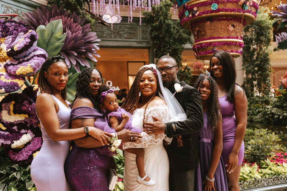 A joyful wedding party poses among lush purple floral arrangements, featuring a bride in white and guests in elegant purple gowns.